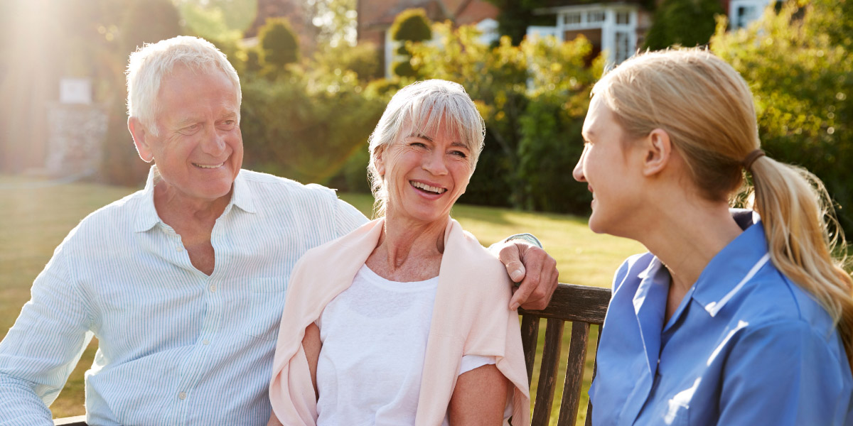 An older couple enjoys a bright afternoon outdoors, smiling and sitting together on a wooden bench in a garden.