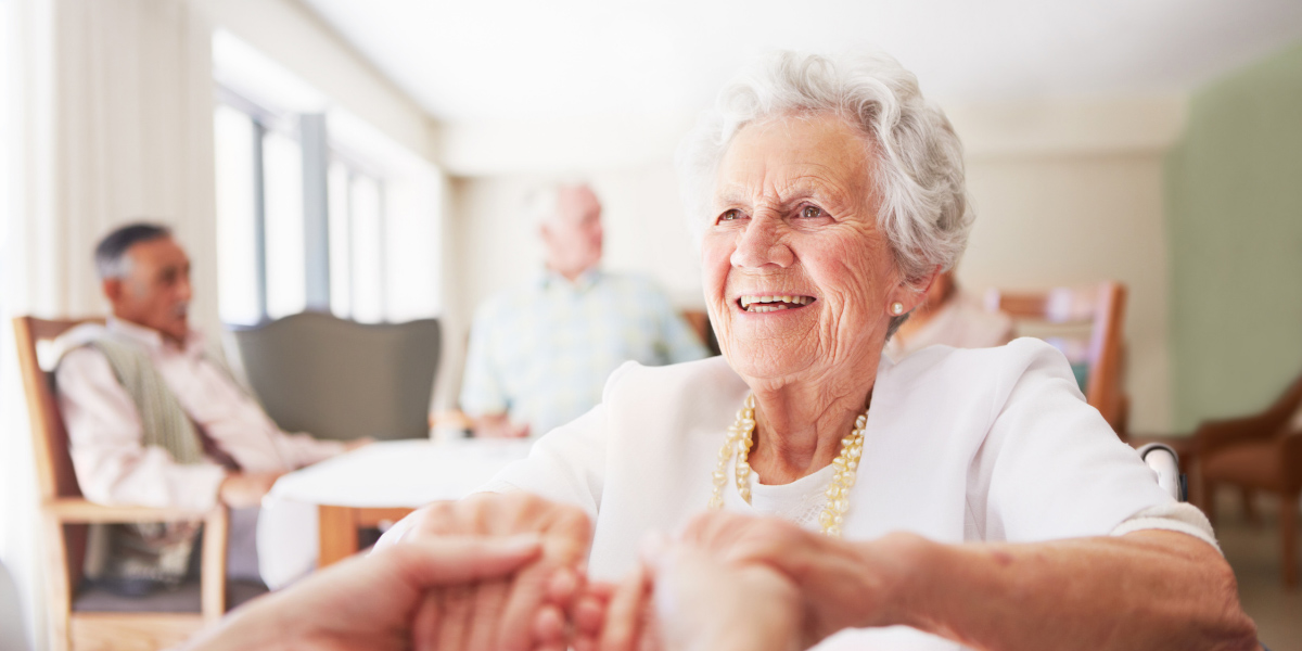A close-up shot of a happy senior woman smiling warmly while holding someone's hands in a bright, indoor room.