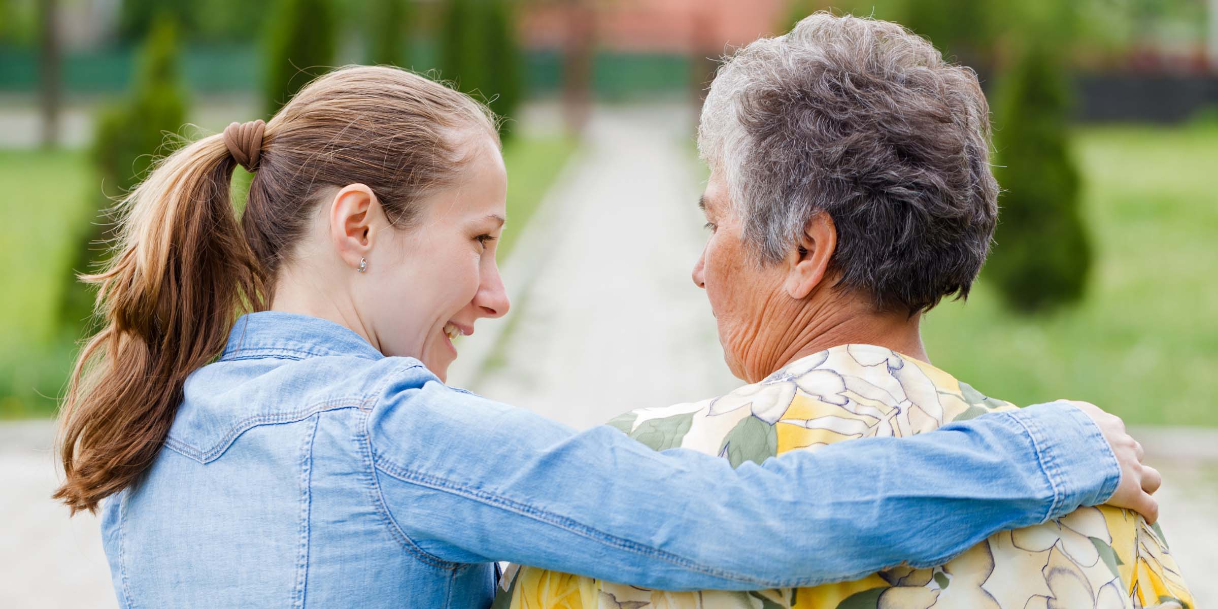 A younger woman in a denim shirt hugs a senior woman from behind while they both smile outdoors.