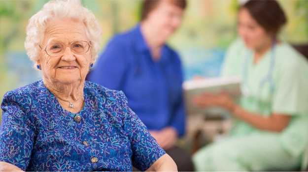 A smiling senior woman in a blue patterned shirt sits in the foreground while others talk in the blurred background.